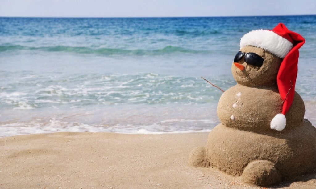 Sand snowman wearing sunglasses and a Santa hat on a sunny winter beach